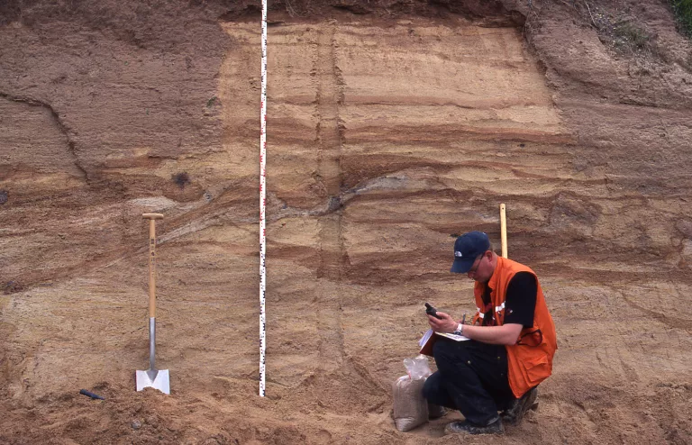 Rotbraune Steinbruchwand, davor ein Geologe in Warnweste und mehreren Arbeitsutensilien
