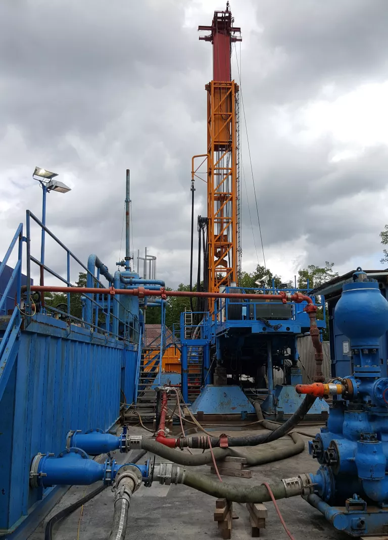 Bohranlage: im Hintergrund ein hoher orangefarbener Bohrturm mit Bohrgestänge aus Metall, im Vordergrund links eine blaue Spülungsanlage, verbunden durch schwarze Schläuche mit Pumpen rechts