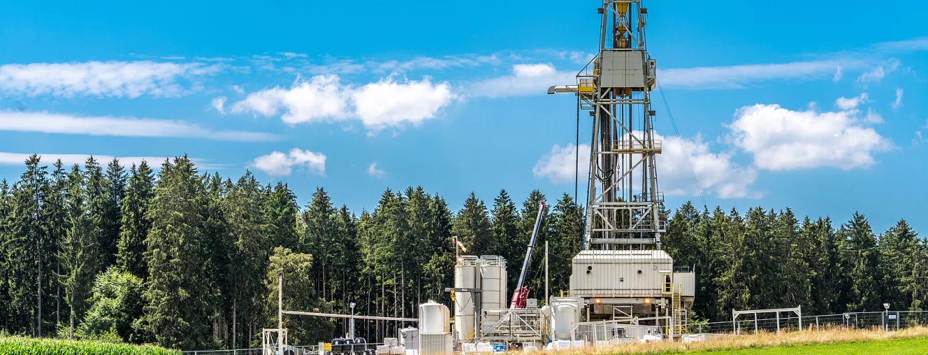 Im Vordergrund eine Wiese, dahinter Wald, im Hintergrund blauer Himmel. Auf der Wiese steht eine Bohranlage mit Metallgitterturm