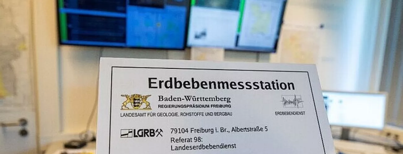 Schild "Erdbebenmessstation" in einem Raum, im Hintergrund mehrere Monitore