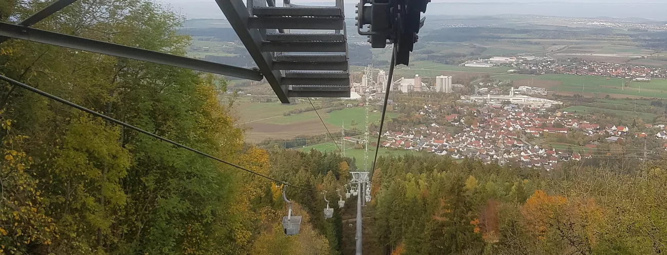 Blick entlang einer Seilbahn nach unten durchLaubwald, im Hintergrund eine Ortschaft und eine Industrieanlage