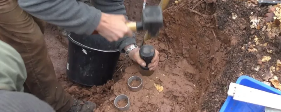 In einer in rötlichem Tonboden aufgegrabenen Profilgrube werden rundliche Metallzylinder eingeschlagen, zur Entnahme von Bodenproben.