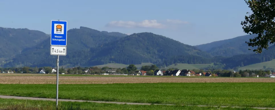 Schild mit Aufschrift "Wasserschutzgebiet". Im Hintergrund Wiesen- und Ackerflächen sowie bewaldete Hänge des Schwarzwalds.