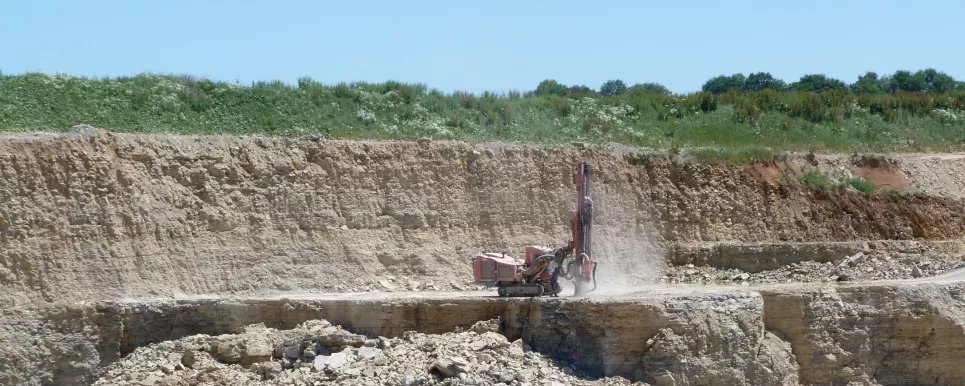 Zu sehen ist ein aktiver Steinbruch in dem ein Bohrgerät, das einem Bagger ähnelt, vertikal eine Bohrung abteuft. 